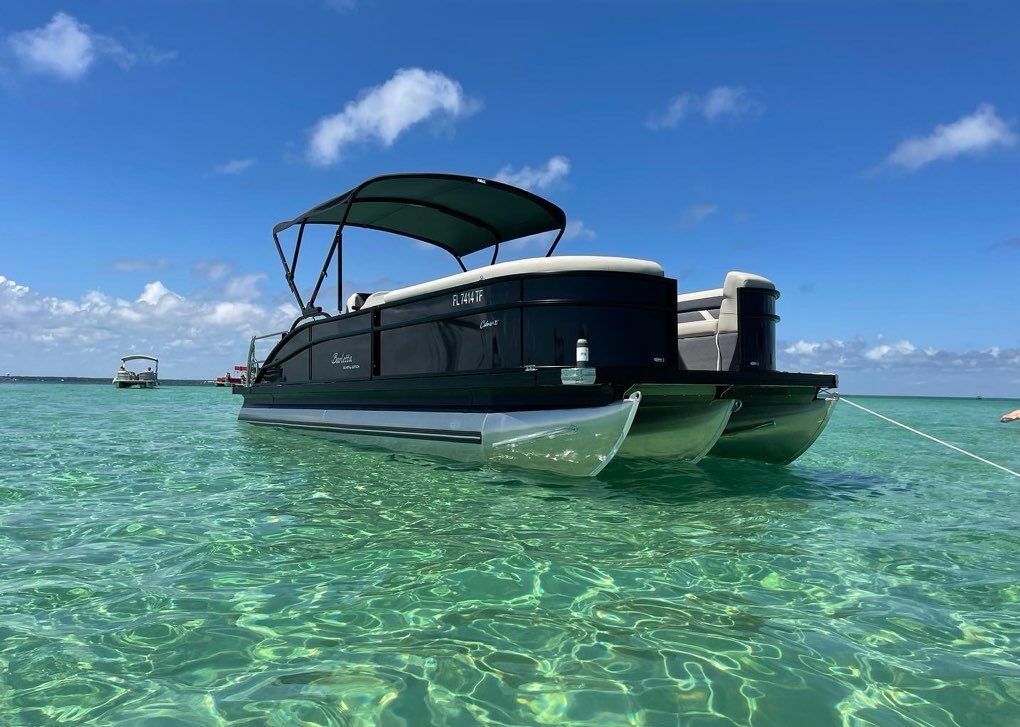 Pontoon boat with sun canopy anchored in shallow crystal-clear turquoise water under a bright blue sky, sunlit ripples and distant boats on the horizon.