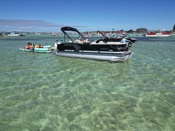 Sunny summer scene of pontoon boats anchored in clear shallow turquoise water with people lounging on inflatable rafts and many boats in the distance at a busy coastal anchorage