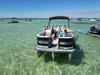 Pontoon boat anchored in shallow crystal-clear turquoise water with people relaxing onboard, other boats nearby and a sunny blue sky — lively sandbar boating scene.