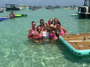Group of women, mostly in pink swimsuits, posing in shallow clear turquoise water beside an inflatable floating dock with anchored boats and beachgoers in the sunny background.