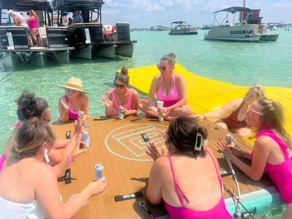 Group of women in pink swimsuits seated in a circle on a floating wooden platform in clear turquoise shallow water, holding canned drinks and chatting with a yellow inflatable mat and anchored pontoon boats in the background — sunny beach party on a calm coastal sandbar.