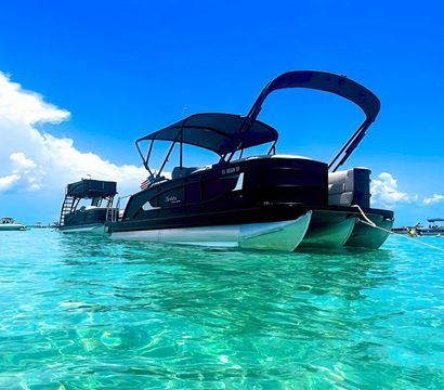 Black pontoon boat anchored in crystal-clear turquoise water under a bright blue sky, with other boats visible in the distance, perfect day for boating.