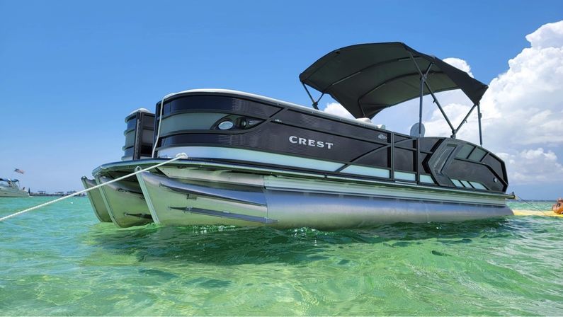 Sleek dark-gray pontoon boat anchored in shallow crystal-clear turquoise water under a bright blue sky with puffy white clouds and a folded bimini top
