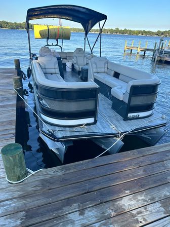 Pontoon boat moored at a wooden lake dock on a sunny day — beige cushioned seating, black bimini canopy, and a yellow foam float strapped to the stern with calm blue water and tree-lined shoreline beyond.