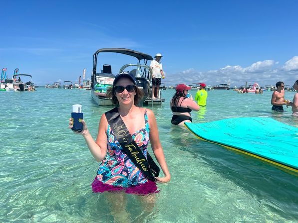 Smiling woman in sunglasses wearing a "50 & Fabulous" sash standing in clear shallow turquoise water near anchored boats, holding a drink while paddleboards and groups of people enjoy a sunny sandbar day.