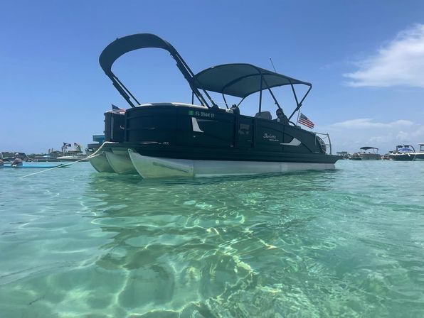 Pontoon boat with canopy and American flag anchored in crystal-clear turquoise shallow water under a sunny blue sky, with other boats visible in the distance.
