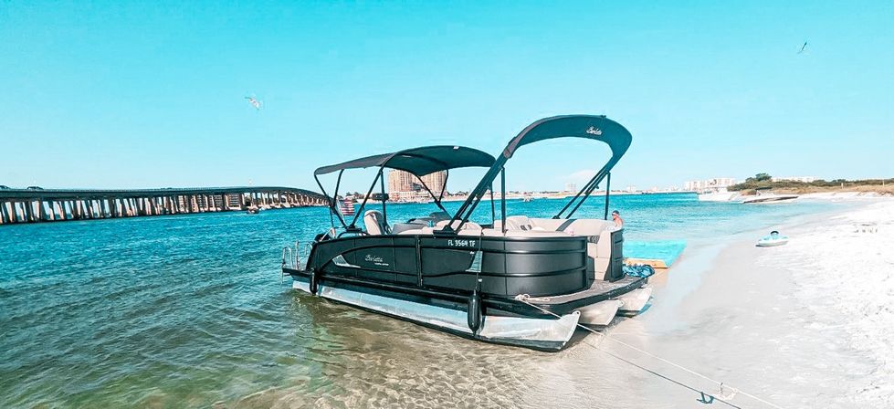 Black pontoon boat beached on a sandy shore with turquoise water, a long bridge in the background and a clear blue sky on a sunny day