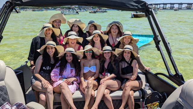 Smiling group of women in wide-brim straw sun hats posing on a pontoon boat during a sunny summer outing in shallow green coastal waters, with other boats and a bridge on the horizon.