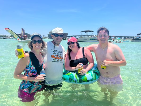 Four adults smiling waist-deep in shallow clear turquoise water on a sunny beach day, wearing swimsuits, hats and sunglasses, holding canned drinks and leaning on a green leaf-patterned inflatable ring with pontoon boats and blue sky in the background.
