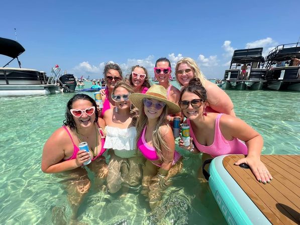 Group of women in pink swimsuits and sunglasses smiling and holding canned drinks while standing in clear turquoise shallow water at a sunny sandbar with pontoon boats in the background