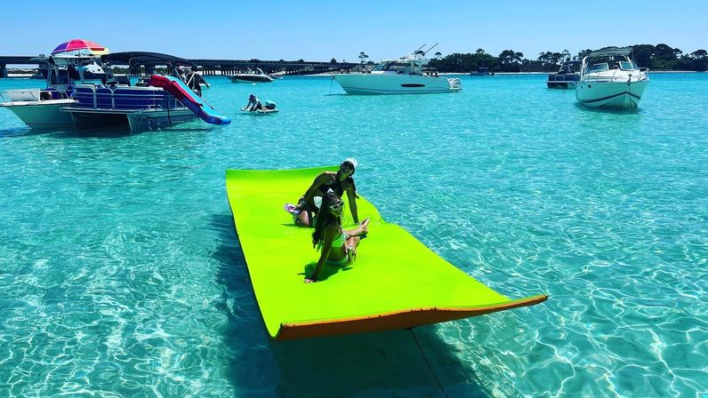 Two people lounging on a large lime-green floating mat in crystal-clear turquoise bay with anchored boats and a sunny blue sky
