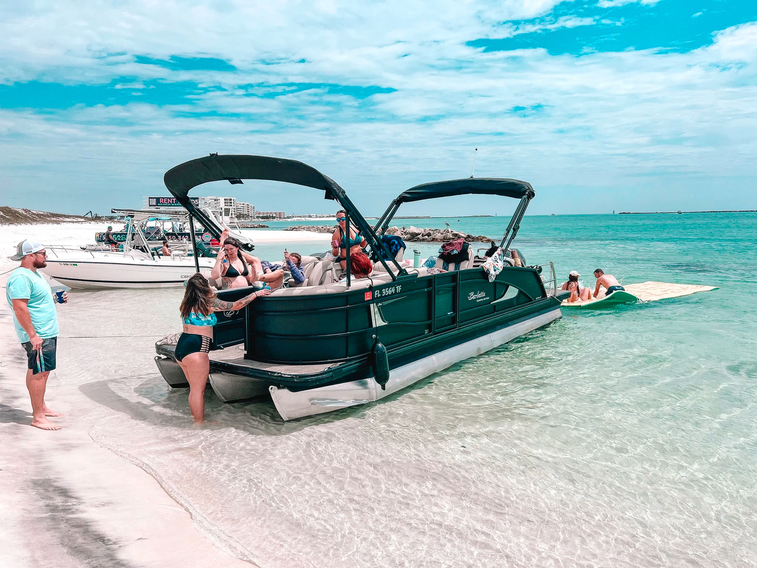 Group of people in swimsuits enjoying a pontoon boat beach party anchored in shallow clear turquoise water off a white sandy shoreline under a bright blue, partly cloudy sky.