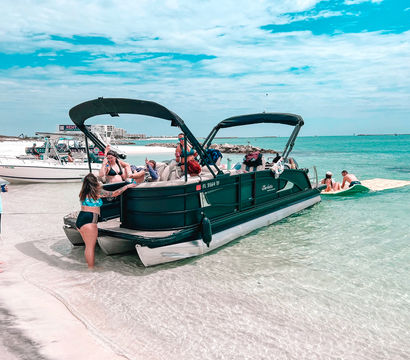 Group of people in swimsuits enjoying a pontoon boat beach party anchored in shallow clear turquoise water off a white sandy shoreline under a bright blue, partly cloudy sky.