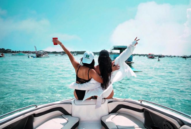 Two friends on the bow of a motorboat with arms raised—one holding a red cup—enjoying clear turquoise coastal waters dotted with other boats on a sunny summer day.