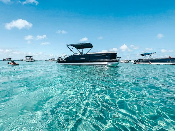 Pontoon boats anchored over crystal-clear turquoise shallow water with people floating nearby under a bright blue sky — tropical boating and beach day vibes.