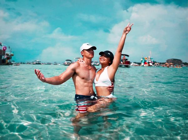 Young couple standing waist-deep in clear turquoise water, woman in white bikini flashing a peace sign, man in cap and striped swim trunks, boats and sunny sky in background.