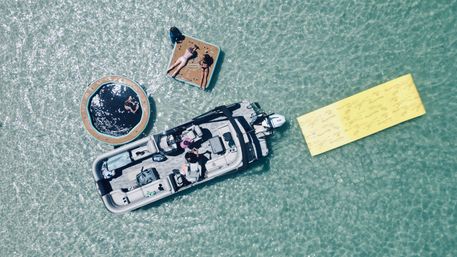 Aerial drone view of a gray pontoon boat anchored in clear turquoise shallow water next to a round inflatable trampoline, a square floating sunbathing platform with loungers, and a long yellow floating mat — summer boat outing.