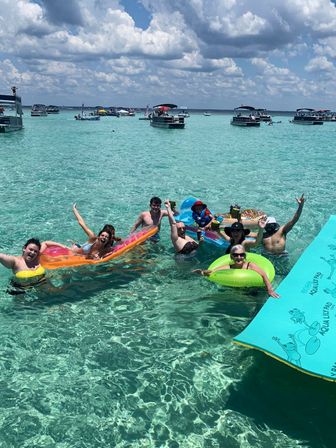 Cheerful group of people on colorful floats in crystal-clear turquoise water near anchored pontoon boats under a sunny blue sky with puffy clouds