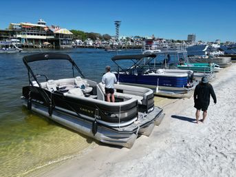 Pontoon boats moored along a sandy beach at a busy waterfront marina on a sunny day, people on decks, colorful waterfront buildings and clear blue sky in the background.