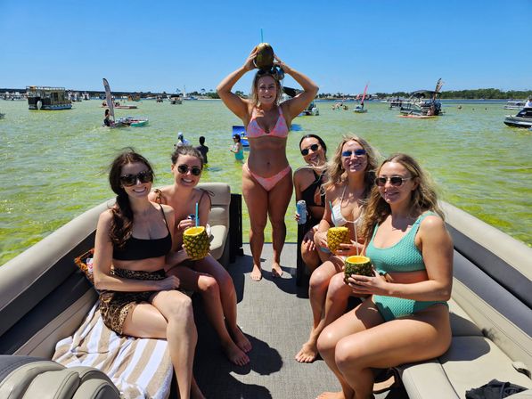 Six women in bikinis on a pontoon boat at a sunny sandbar—summer party sipping pineapple and coconut drinks with many boats on shallow green water.