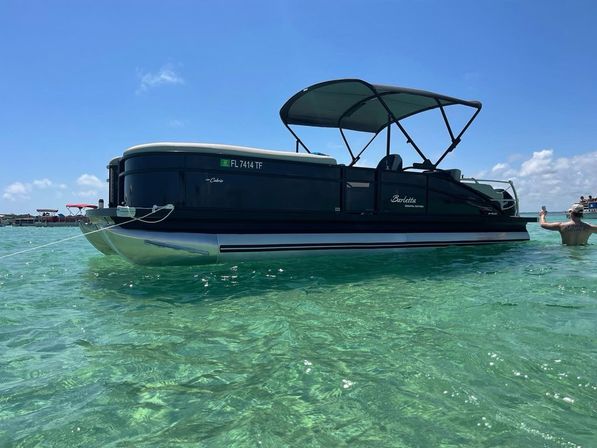 Dark pontoon boat with canopy floating in clear turquoise Florida waters on a sunny day, with people wading nearby.