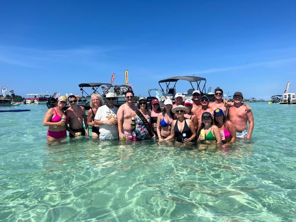 Group of people posing in waist‑deep clear turquoise water at a sunny sandbar, holding drinks with pontoon boats, colorful flags and a bright blue sky in the background.
