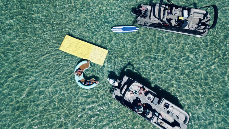 Aerial view of two pontoon boats anchored in crystal-clear turquoise shallows with people relaxing on a C-shaped inflatable lounger, a yellow floating mat and a blue paddleboard — sunny summer boating scene.