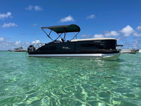 Black pontoon boat with canvas canopy anchored in shallow crystal-clear turquoise water under a sunny blue sky with distant boats on the horizon