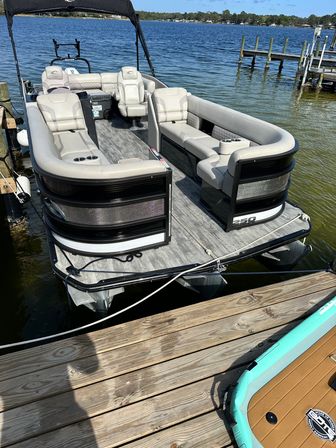 Pontoon boat tied to a wooden dock on a sunny lake — spacious gray cushioned seating with cup holders, open deck, turquoise paddleboard at the dock, calm blue water and tree-lined shore.