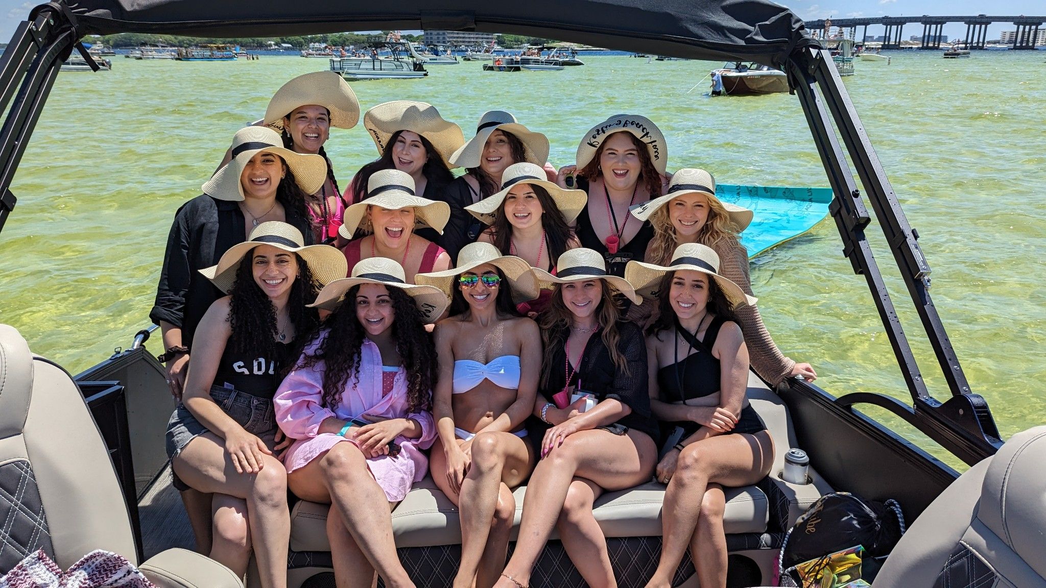 Smiling group of women wearing wide-brim sun hats on a pontoon boat for a sunny bachelorette-style outing in clear green bay waters, with other boats and a bridge visible in the background.