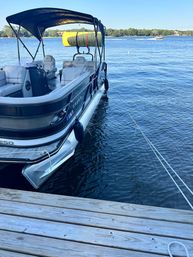Pontoon boat with black canopy and cushioned seats docked at a wooden pier on a calm blue lake, yellow paddleboard on the rack and a speedboat in the distance.