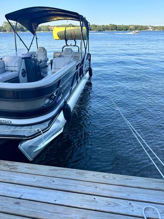 Pontoon boat with black canopy and cushioned seats docked at a wooden pier on a calm blue lake, yellow paddleboard on the rack and a speedboat in the distance.