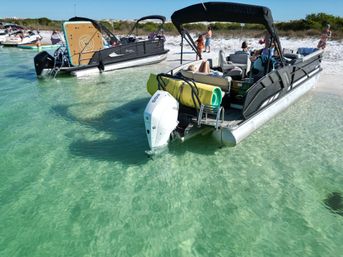 Pontoon boats anchored in shallow clear turquoise water off a sandy beach on a sunny day, people lounging aboard and on the shore for a relaxed coastal outing.