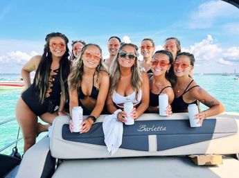 Group of smiling women in swimsuits wearing matching orange sunglasses and holding canned drinks, posing on a boat over clear turquoise water under a sunny blue sky.
