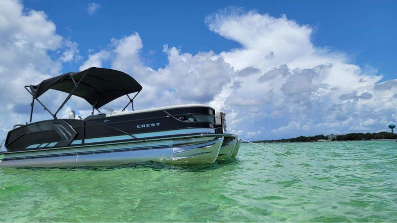 Pontoon boat with a black canopy anchored in clear turquoise water, sunlit emerald waves beneath a bright blue sky with fluffy cumulus clouds and a tree-lined shoreline in the distance.