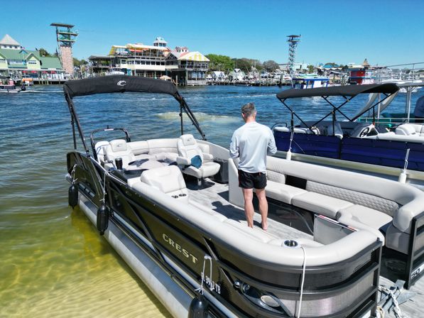 Person standing on a luxury pontoon boat docked at a sunny marina with waterfront restaurants, neighboring boats, and a clear blue sky.