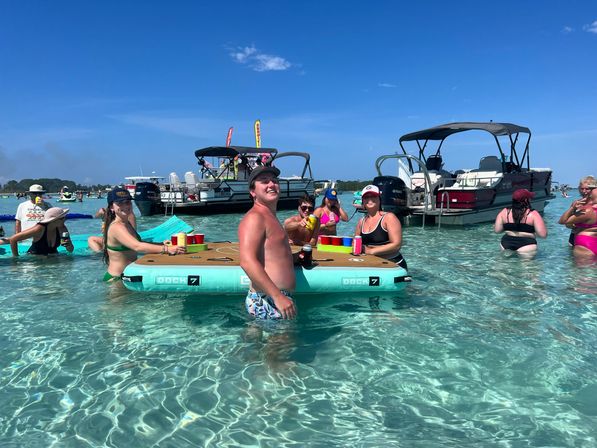 People playing a floating beer‑pong game in crystal‑clear shallow turquoise water near anchored pontoon boats on a sunny coastal day, colorful cups and swimsuits creating a festive beach‑party vibe.