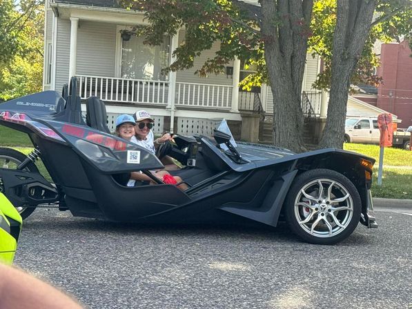 Black three-wheeled open-top sport trike with two smiling passengers wearing caps, parked on a tree-lined residential street in front of a porch house.