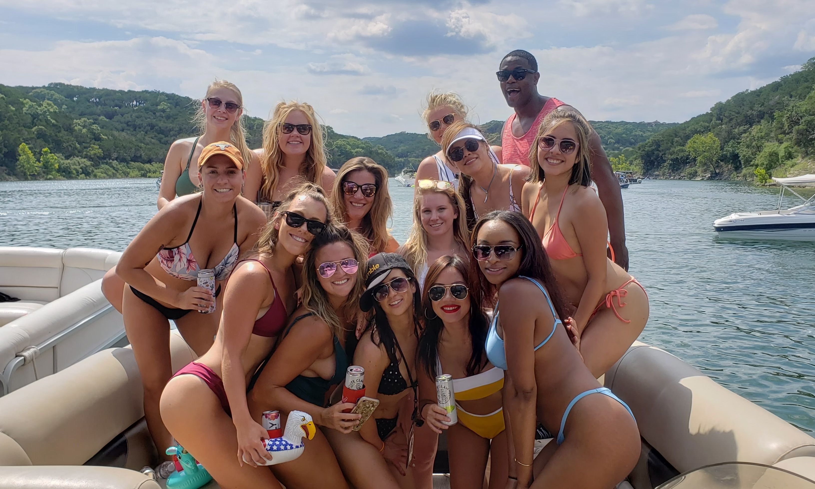 Smiling group of young adults in colorful swimsuits posing on a pontoon boat with drinks on a sunny, tree-lined lake — summer boating party