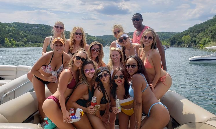 Smiling group of young adults in colorful swimsuits posing on a pontoon boat with drinks on a sunny, tree-lined lake — summer boating party