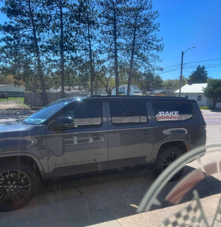 Dark gray SUV parked on a sunny suburban street with tall pine trees, single-story houses and a bright blue sky, rear window showing a small decal and reflections in the glass.