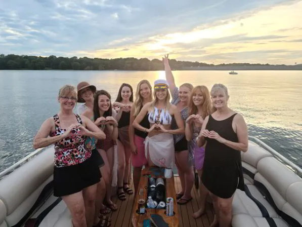 Cheerful group of women posing on a pontoon boat at sunset over a calm lake, many making heart hand gestures and one wearing a captain's hat.