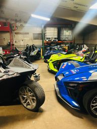 Row of colorful three-wheeled Polaris Slingshot roadsters (blue, neon yellow, silver) parked inside a cluttered workshop garage with shelving and storage tanks.