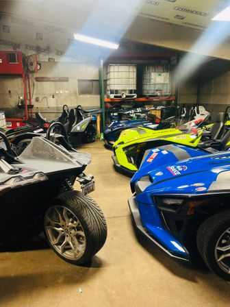 Row of colorful three-wheeled Polaris Slingshot roadsters (blue, neon yellow, silver) parked inside a cluttered workshop garage with shelving and storage tanks.