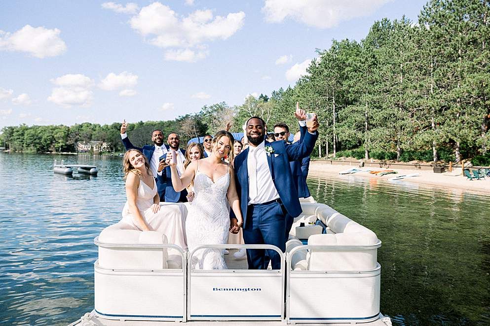 Newlyweds and bridal party cheering aboard a pontoon boat on a sunny lake, holding drinks with smiling friends and a forested shoreline and sandy beach behind them.