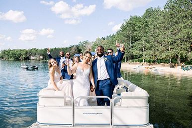 Newlyweds and bridal party cheering aboard a pontoon boat on a sunny lake, holding drinks with smiling friends and a forested shoreline and sandy beach behind them.