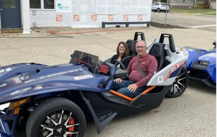 Two people smiling while seated in a blue three-wheeled open-top sports roadster with orange trim, parked on a city street in front of a building under construction.