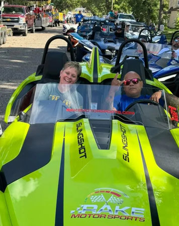 Lime-green open-top three-wheeled roadster with two riders smiling and gesturing, lined up with other vehicles in a sunny neighborhood parade on a tree-lined street