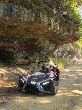 Sleek three-wheeled open-air roadster parked on a sunlit dirt road beneath a graffiti-covered limestone rock overhang with trees and rugged cliffs in the background