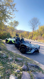 Two sporty three-wheeled open-top roadsters parked on a scenic country road amid fall foliage and clear blue sky